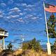 Double Arrow Lookout Tower- Tower in background, flag pole with American flag in foreground, fluffy grey clouds pass over a partly cloudy blue sky