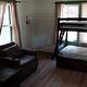 Bunk beds and chairs in the main living room seen from the kitchen. The scene is dimly lit as sunlight enters the room through 3 large windows.