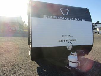 Photograph of a travel trailer parked on a gravel lot with the dealership buildings and outdoor display area visible — documents the lot and typical vehicles on the property (from the dealership listings).