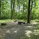 A open campsite with fire grill, picnic table, and rock barriers in the foreground and green vegatation background. 