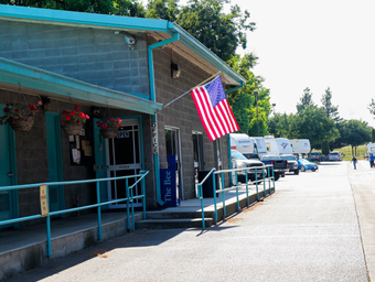 Shows the RV Park office/building with an American flag and a line of RVs parked along a paved drive — an on-site facility image consistent with the RV Park page background.