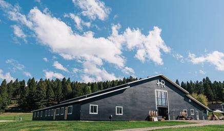Wide exterior shot of the large black barn/event building on the property with grassy foreground and treeline — clearly depicts the main facility and setting.