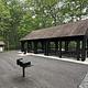 Opened sided, roofed picnic pavilion in the forest near a flat surface with two standing grills with a restroom building nearby