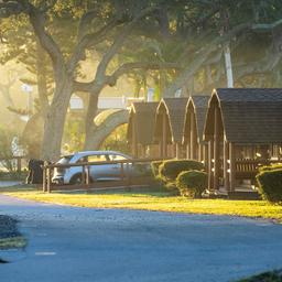 Row of KOA-style cabins along a roadway with mature trees and parked car — shows cabin exterior layout and campground lanes at sunrise. (waystostay photos).