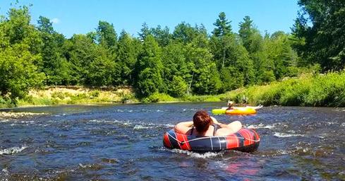 People tubing on a river running through wooded parkland — matches amenities description of river/kayak/tubing (page: https://www.pineridgeparkcampsite.com/amenities). Shows recreational water feature on the property.