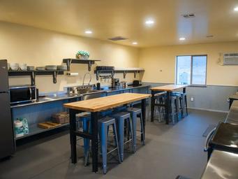 Communal kitchen with stainless steel counter, dish shelves, microwave, and butcher-block high tables with blue metal stools.