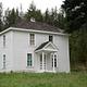 White, two-story house on a grassy hill next to a fir forest.