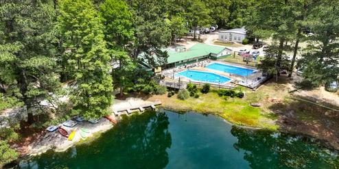 Drone/oblique view focused on the pond, pool complex and docks with kayaks — highlights amenities and waterfront features on the campground grounds.