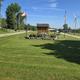Clear photo of the campsite entrance/central lawn with flags, a sign kiosk, access road and RVs visible in the distance — matches the Home page context showing property grounds and entry.