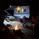 Nighttime campsite scene with a travel trailer, picnic table and campfire with people — illustrates evening camping atmosphere and fire-ring facilities shown on the campground’s pages.