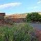 A paved road. To the right side there is desert fauna and a brown sign reading: "Loop C *right arrow* Amphitheater *right arrow* Group Area *right arrow*". To the left side is a fence and some trees. There is a brown-red color hill against a blue sky in the background.