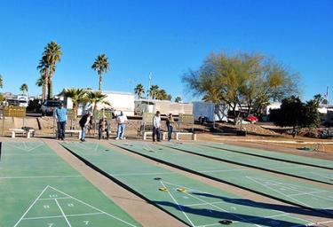 Wide view of outdoor shuffleboard courts with people playing and RVs/landscaping in background — documents on-site recreational amenities visible on the resort page. 