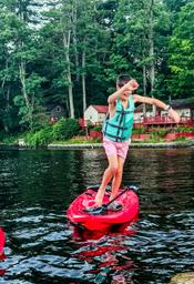 A young boy wearing a turquoise life vest and pink shorts balancing on a red paddleboard on the river, with trees and cabins in the background.