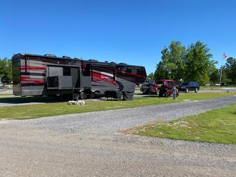 Wide daytime shot of a large motorhome parked on a grassy, gravel site with utility pedestals and neighboring sites visible — good example of 'big rig friendly' RV accommodations.