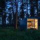 Twilight/night shot of a small modern cabin with interior lights visible through large windows set in the woods — offers an atmospheric view of the property’s lodging experience.