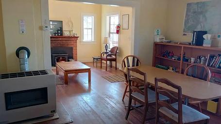 View of living room and dining room. Table has 4 chairs (more in storage), shelf equipped with First Aid kit, history/diary booklets, toys/games, and miscellaneous books.