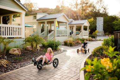 Depicts small cabin units and paved paths with guests (kids riding carts) — a clear photo of the campground's cabin area; sourced from the property 'waystostayphotos' gallery on the KOA campsite page.
