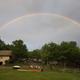 Hero photo showing wide view of the campground: grassy foreground, playground equipment, RVs and buildings among trees with a full rainbow overhead — good overall overview of site from the campground’s official page (social/hero image).