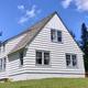 White house with dormer roof on a grassy hill with conifer trees.
