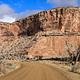 A dirt road cuts through a wide canyon of orange cliffs with a bathroom in the distance.