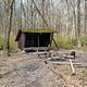 Three-sided wooden Adirondack shelter, 2 wooden benches and fire circle.  Surrounded by trees with no leaves.