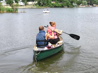 Two people canoeing on a lake with shoreline and RVs visible in the distance — depicts canoeing activity and the campground’s lake recreation (from site gallery/feature images).