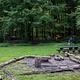 Metal fire ring/grill surrounded by logs and picnic tables in open field.  Multiple tents visible in the distant woods.