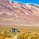 Gray campervan on roadside campsite with green brush vegetation in the foreground and dramatic reddish and white stratified mountains in the far background.