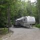 Gravel RV site with a fifth-wheel camper and awning in a wooded setting — good close-up of typical RV pitch, from Amenities page.