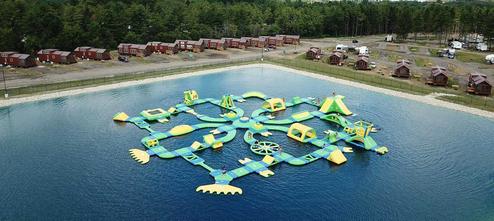Aerial/oblique shot showing lake inflatables with a row of cabins or park-models along the shore — highlights lodging/rental units and water feature on the property (campgrounds-main_0069).