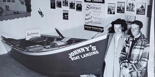 A vintage black and white photo of two people standing beside a boat labeled 'Johnny's Boat Landing'.