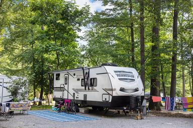 Shows an RV parked at a gravel, wooded campsite with outdoor setup — clear depiction of RV site layout and utilities. Matches on‑site 'RV Sites' imagery in the property gallery.