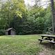 Green grass field with a wooden picnic table on the right and an outdoor toilet on the left
