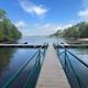 A wooden dock extending into a calm lake with floating boat slips, surrounded by green trees under a partly cloudy blue sky.