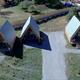 Aerial view of three small wooden A-frame cabins with metal roofs and covered porches, set along a gravel driveway on a grassy lot.
