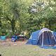 A blue tent sits on a campsite with a thick row of green trees behind the tent.