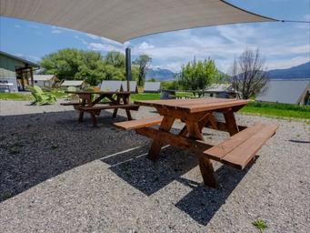 Wooden picnic tables on a gravel patio under a beige shade sail, with green lawn, trees, and snow-capped mountains beyond.
