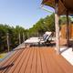 Wooden deck with two lounge chairs under a canvas canopy, overlooking lush green trees against a blue sky.