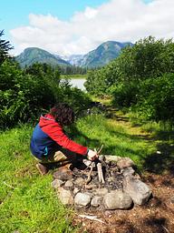 A person building a teepee of sticks in a rock firepit with mountains is background
