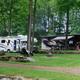 Multiple RVs parked among trees with a central firepit and stonework — illustrates campsite/RV pitch layout and communal fire area (About/Rates pages).