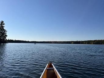 The front of a canoe surrounded by a blue lake with another canoe in the distance.