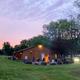 Wide outdoor shot at dusk showing a lodge/building, grassy lawn and a central campfire ring with people — good view of campground social area and facilities.