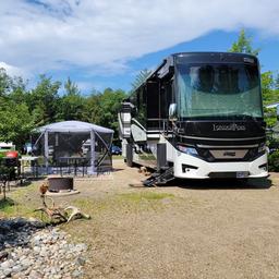 Wide view of a large RV parked at a leveled gravel/KOA-style site with patio, fire ring and picnic setup — concrete evidence of RV accommodations on the property.