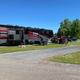 Wide daytime shot of a large motorhome parked on a grassy, gravel site with utility pedestals and neighboring sites visible — good example of 'big rig friendly' RV accommodations.