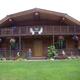 Clear daytime exterior of a large log cabin on manicured lawn with porch/upper balcony and hanging flower baskets — matches Cloudberry Cabins homepage hero image and site context showing on‑site cabins.