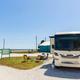 A white RV is parked at a campground near the beach with two empty chairs and a small table set up outside on a sunny day.