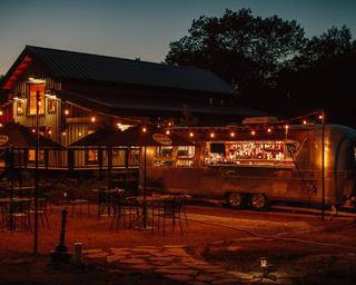 Evening shot of an Airstream bar and lit outdoor seating in front of a building — illustrates on‑site food/beverage service and nighttime atmosphere.
