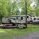 Pull-through RV site photo showing a fifth-wheel parked on a wooded, level site with landscaping and gravel access — useful to show RV accommodations (from waystostay photos on the campground page).