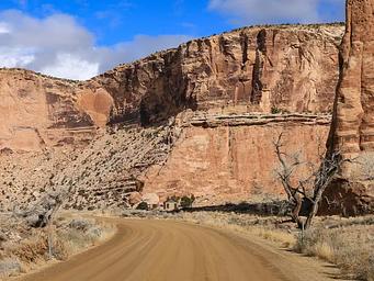 A dirt road cuts through a wide canyon of orange cliffs with a bathroom in the distance.