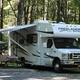 Class C motorhome at a shaded RV site with picnic table and awning — clear depiction of RV camping spaces and site setup from the homepage images.